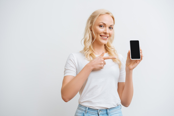 happy woman pointing at smartphone with blank screen while looking at camera isolated on white - Fotó, kép