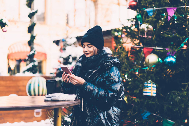 Thoughtful trendy young woman in warm clothes using smartphone and drinking coffee near adorned Christmas fir tree on blurred background  - Fotó, kép