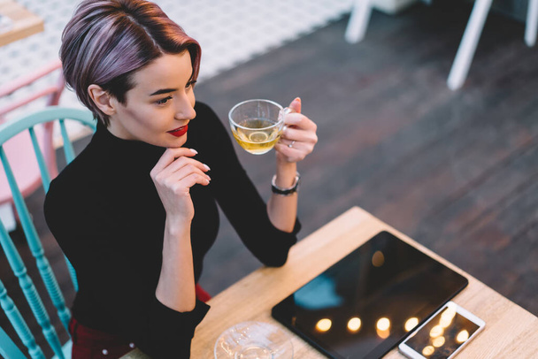 From above stylish content woman with short hair in casual clothes drinking cup of tea while sitting at table with smartphone and tablet at hipster cafeteria - Foto, immagini