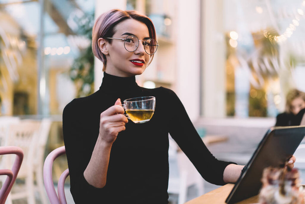 Stylish happy woman with short hair in casual clothes and glasses drinking cup of tea and using tablet while sitting at table at hipster cafeteria - Foto, imagen