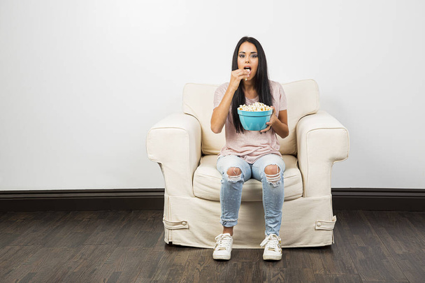 Young woman sitting on a white couch, eating popcorn from a blue bowl - Foto, afbeelding