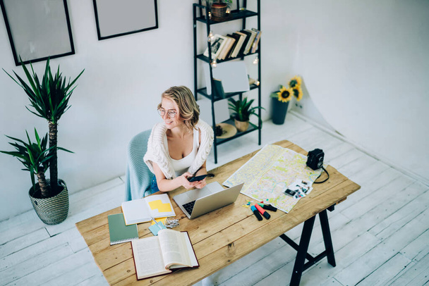 Top view of happy female freelancer with smartphone device in hand sitting at comfortable work place for distance job looking away and smiling while planning travel vacations, self employed woman - Foto, immagini
