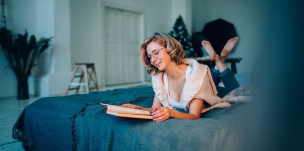 Cheerful beautiful blonde woman in casual clothing and glasses reading book while lying on comfy bed in modern studio room - Foto, immagini
