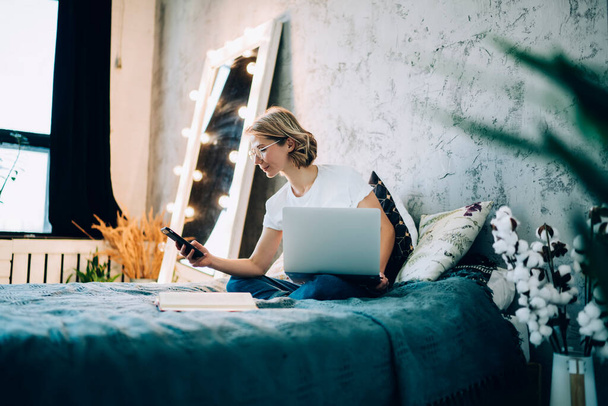 Attractive Caucasian female blogger taking rest in home apartment using 4g wireless for checking smartphone notification during time for watching movie via laptop computer, skilled woman on bed - Foto, Imagem