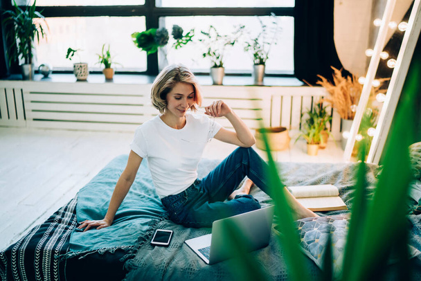 Delighted focused attractive woman recreating on cozy soft bed while using laptop and smartphone and reading absorbing book in loft  - Fotografie, Obrázek