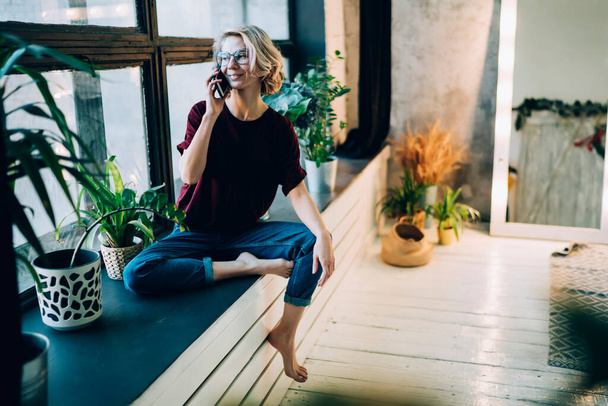 Charming smiling positive young barefoot woman in casual outfit with glasses speaking on smartphone while sitting on windowsill beside plants in pots on evening - Φωτογραφία, εικόνα