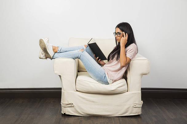20 και κάτι γυναίκα, κάθεται σε ένα λευκό καναπέ, διαβάζοντας ένα σκληρό εξώφυλλο και προσαρμόζοντας τα γυαλιά της - Φωτογραφία, εικόνα