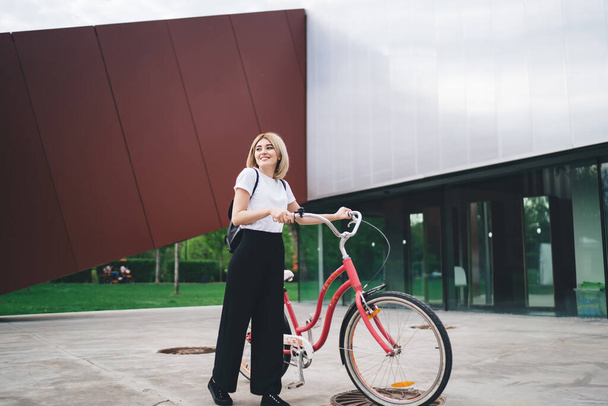Happy and cheerful lady in casual clothes walking near modern building with red bicycle and smiling while looking at camera  - Valokuva, kuva