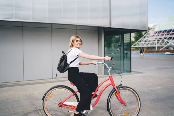 Seitenansicht der jungen attraktiven Studentin in lässiger Kleidung mit Rucksack sitzt auf einem Fahrrad mit rotem Rahmen und schaut in die Kamera während der Fahrt auf der Straße - Foto, Bild