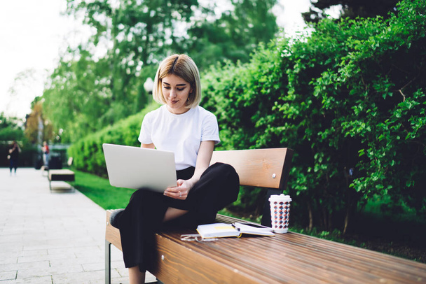Concentrated focused hipster woman in casual outfit alone on wooden bench with notepad and coffee cup in park working remotely on laptop - Фото, изображение