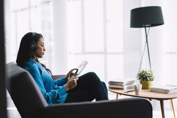 Side view of carefree ethnic woman with headphones sitting with crossed legs in front of coffee table in office and browsing tablet - Foto, Imagem