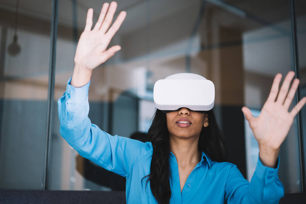 Dark haired excited African American woman experiencing virtual reality using white modern headset and trying to touch imaginary things in air indoors - Foto, immagini