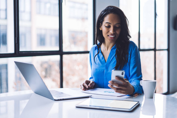 Positive African American woman in formal shirt texting smartphone while sitting by desk with laptop and tablet with empty screen in modern office - Valokuva, kuva