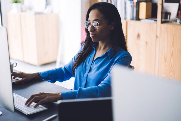 African American businesswoman with slight smile wearing eyeglasses and blue shirt sitting at desk in office and browsing internet on computer with large monitor - Foto, Bild