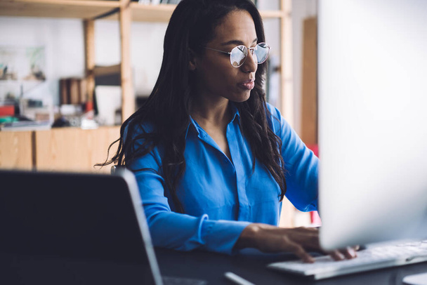 Crop serious focused African American businesswoman in blue shirt and glasses sitting at desk in front of large monitor and typing on keyboard - Foto, Bild