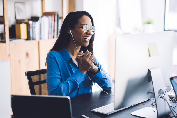 Happy young African American female office worker in blue shirt and glasses sitting at work table with  hands together wearing headphone and conducting online negotiations with coworkers - Zdjęcie, obraz