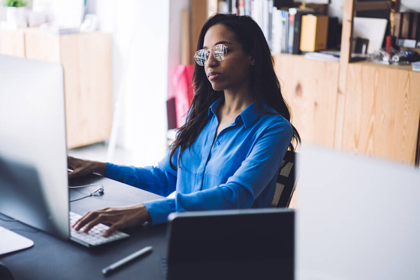 Shora zaostřená mladá Afroameričanka v módních brýlích pomocí klávesnice pro každodenní kancelářskou aktivitu sedící u pracovního stolu - Fotografie, Obrázek