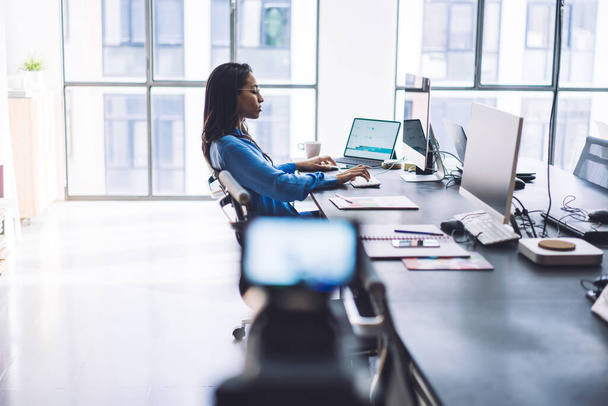 Side view of tired dreaming black businesswoman making film on camera with tripod about work life while using modern devices at office table - 写真・画像
