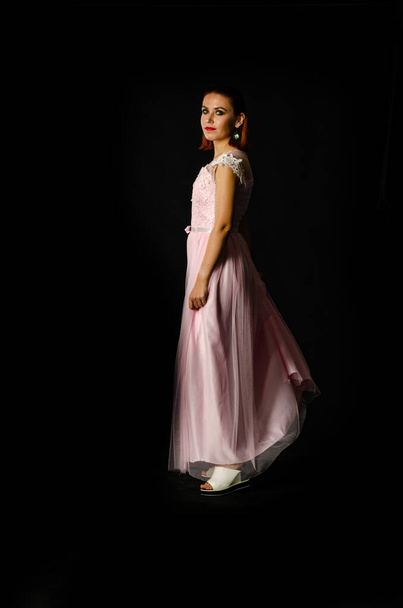 ピンクの長いドレスを着た美しい女性が暗いスタジオの背景にポーズをとって  - 写真・画像