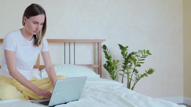 Woman working on a laptop sitting on the bed in the house. - Кадри, відео