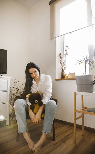 brunette in a white shirt holds a brown bear toy in a stylish interior - Foto, Bild