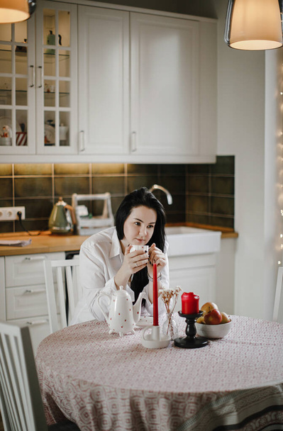 lovely brunette girl in a white shirt drinks tea at a table, kitchen interior design - Foto, Imagen
