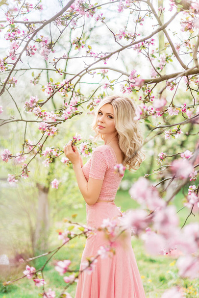 gyönyörű szőke nő egy rózsaszín hosszú ruha egy tavaszi virágzó kertben - Fotó, kép
