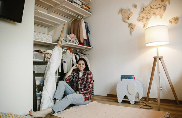 cute pensive brunette in a plaid shirt sits on the floor in the children's room near the wardrobe, wooden elephant and lamp - Foto, Bild