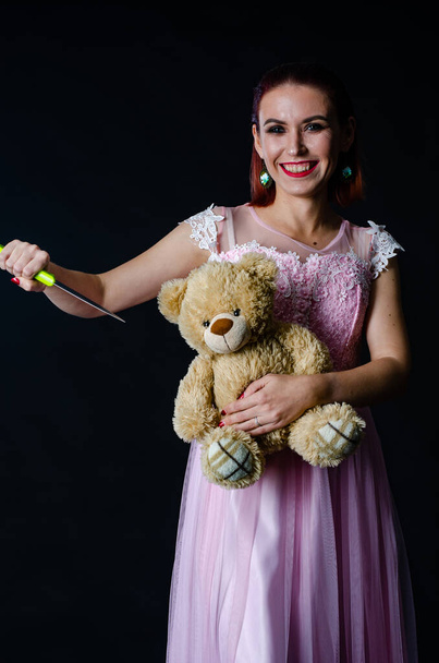 Hermosa mujer pelirroja en lindo vestido rosa sosteniendo oso de juguete y cuchillo en las manos, concepto de Halloween  - Foto, Imagen