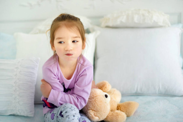 Porträt eines kleinen 3-jährigen schmollenden kapriziösen Mädchens, das im Schlafanzug mit ihrem Spielzeugbär im Bett sitzt. Emotionales Kind. Kind in schlechter Laune. Trauriges kleines Mädchen in Nachtwäsche. - Foto, Bild