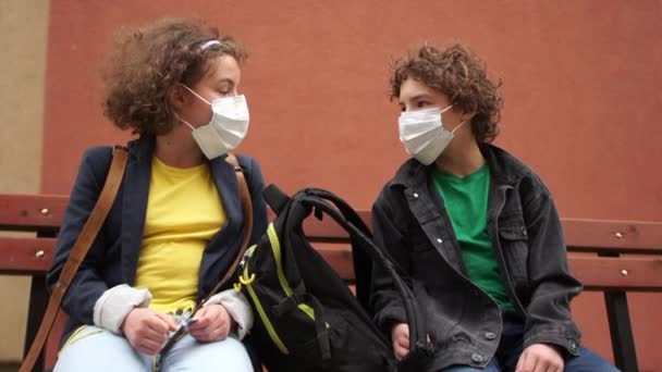 Back to school after quarantine. Cute curly boy and girl are sitting on a bench at a safe distance and talking. Children say goodbye contactlessly, touching their elbows - Filmati, video