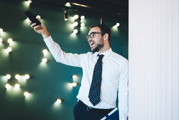 Vigorous happy man in glasses in white shirt and tie putting hand with smartphone up leaning on white wall opposite of green wall with lamps   - Valokuva, kuva