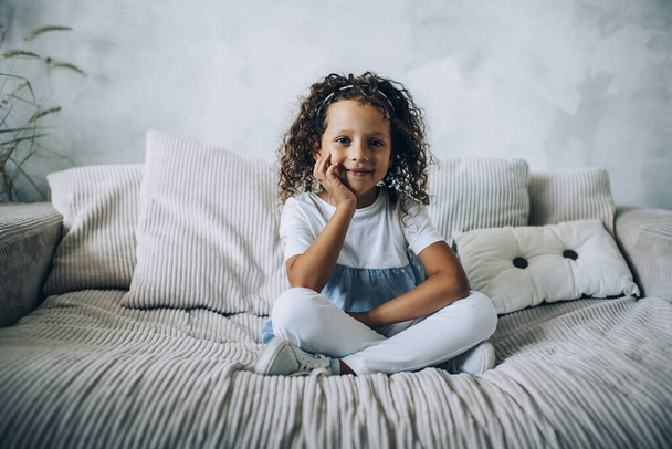 Adorable smiling little kid with curly hair leaning with chin on hand while relaxing on comfortable sofa with cushions looking at camera - 写真・画像