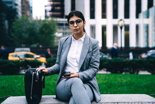 Portrait of intelligent businesswoman in formal wear sitting at city bench and looking at camera, Hispanic lawyer in optical eyeglasses for vision protection holding cellphone for digital marketing - Zdjęcie, obraz