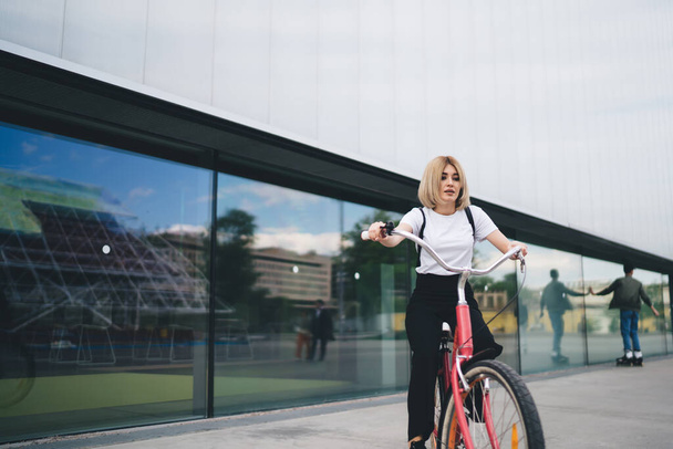 Beautiful trendy casual blond female with backpack riding vintage looking pink bicycle against modern city building in daylight looking down - Photo, image