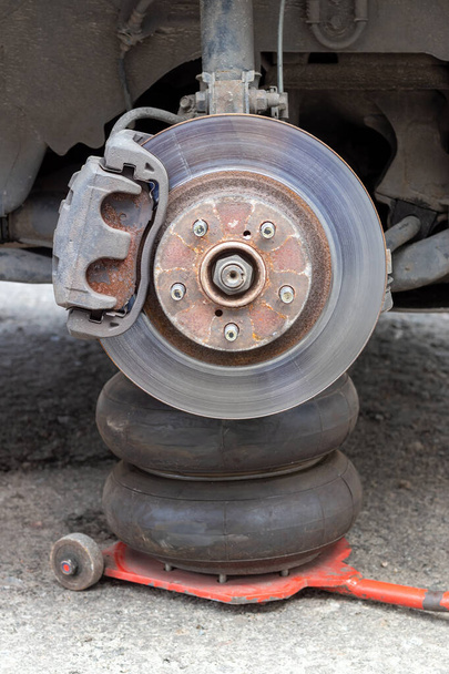 Cubo de rueda de coche oxidado con disco de freno en la tienda de neumáticos. Coche sin rueda levantado en el gato neumático mientras se reemplaza en el centro de servicio de neumáticos. Vista de cerca
 - Foto, imagen