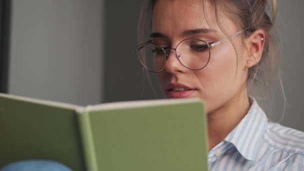A close-up view of a dreaming young woman wearing glasses is making notes in her notebook sitting on the sofa at home - Metraje, vídeo