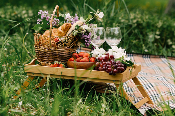 Cesta de piquenique com morangos, uvas e pães na grama verde no jardim
 - Foto, Imagem