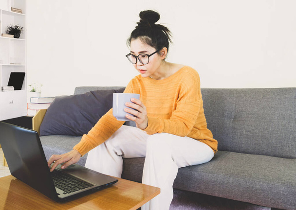 Young beautiful asian woman wore orange knitwear, sitting on gray sofa ,work online lifestyle,relax at home with coffee mug,home working laptop,work from home - Фото, зображення