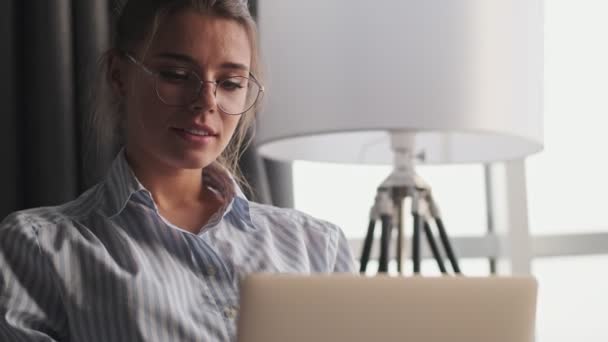 A pretty nice young woman wearing glasses is looking to her laptop sitting on the sofa at home - Πλάνα, βίντεο