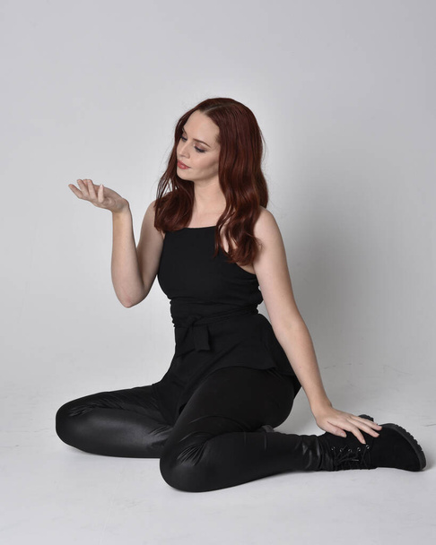 Portrait d'une jolie fille aux cheveux roux portant un pantalon, une chemise et des bottes en cuir noir. pose assise pleine longueur, isolée sur fond de studio
 - Photo, image