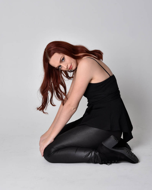 Portrait of a pretty girl with red hair wearing black leather pants, shirt and boots.  full length sitting pose, isolated against a studio background - Фото, зображення