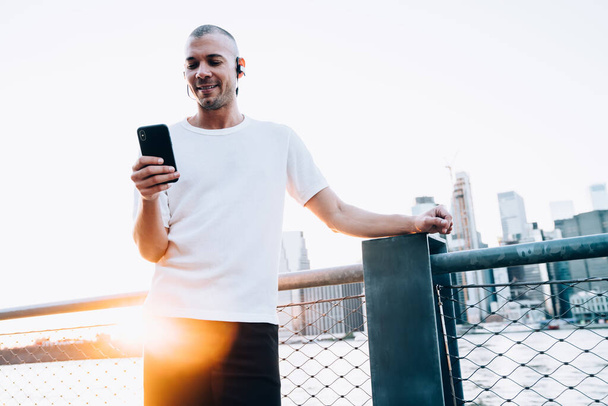Young ethnic smiling short haired man in white shirt with earphones using smartphone and standing near metallic fence and river against background of city - Foto, Imagem