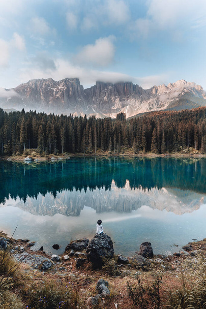 Back view of happy blonde girl against blue sky at Lake Karersee, Dolomites, Italy. Carefree woman with floral dress and a hat enjoys the lake view surrounded by mountains - 写真・画像