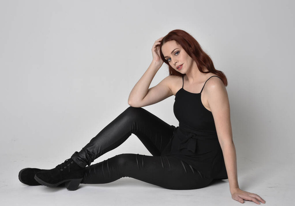 Portrait of a pretty girl with red hair wearing black leather pants, shirt and boots.  full length sitting pose, isolated against a studio background - Φωτογραφία, εικόνα