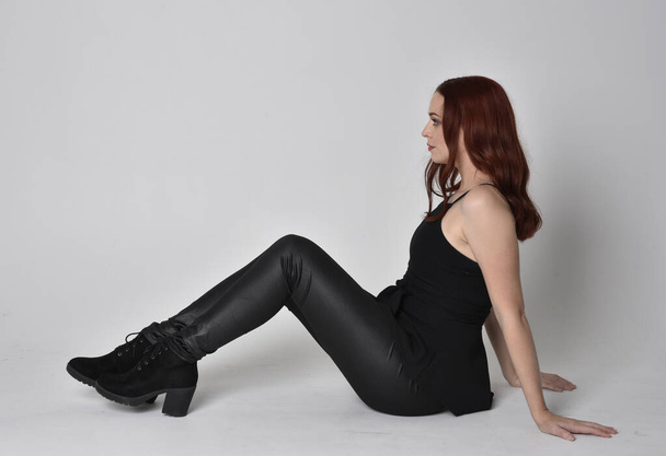 Portrait of a pretty girl with red hair wearing black leather pants, shirt and boots.  full length sitting pose, isolated against a studio background - Foto, Bild