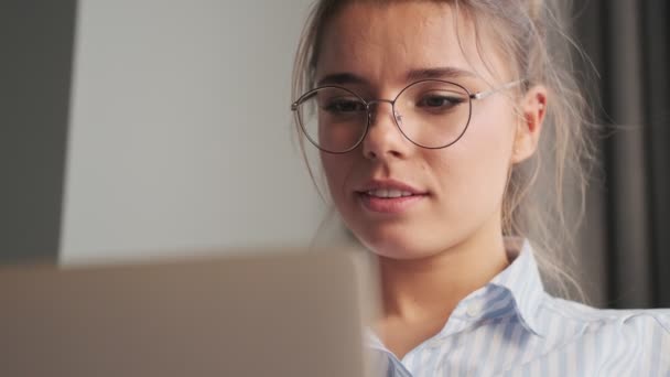 A close-up view of a smiling successful young woman wearing glasses is using her laptop while sitting in the living room at home - Séquence, vidéo