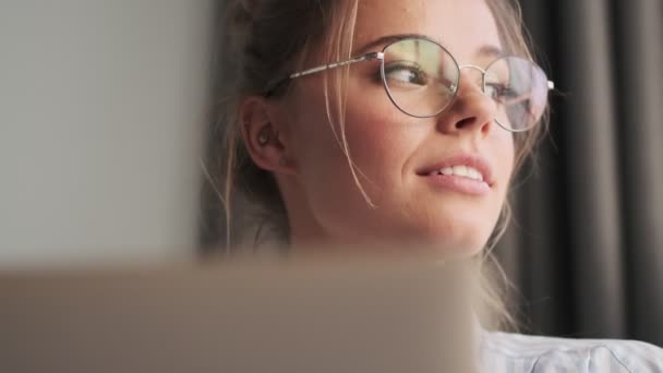 A close-up view of a beautiful dreaming young woman wearing glasses is using her laptop ad looking to the side while sitting in the living room at home - Footage, Video