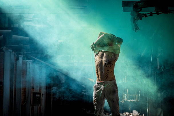 strong man against blue mist at factory exhausted takes off t-shirt, press and side muscles of belly illuminated by light - Fotografie, Obrázek