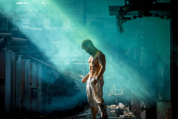 strong man against blue fog at factory exhausted stands, press and lateral abdominal muscles and pectoral muscles illuminated by light, place for text - Foto, immagini